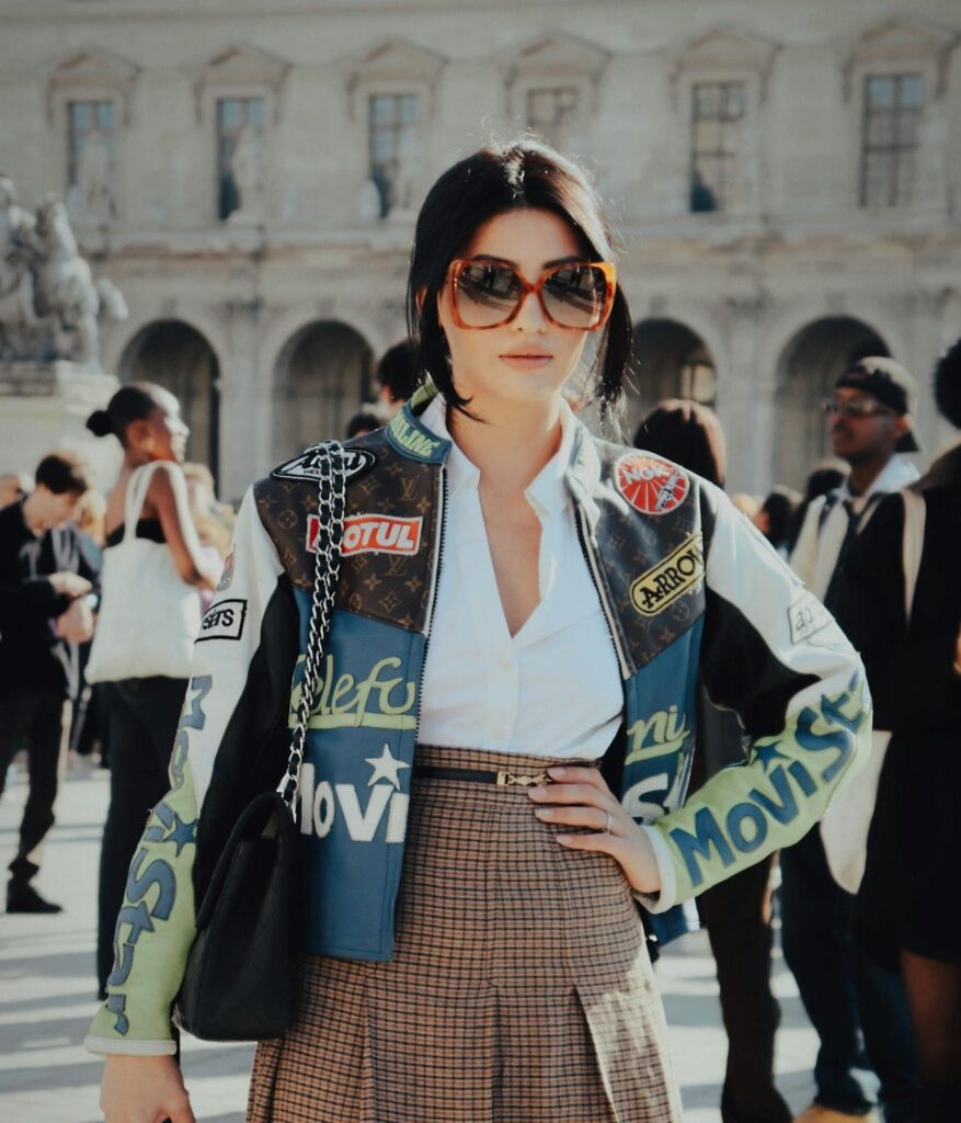 Woman wearing a patterned jacket in front of a historic building, surrounded by people.